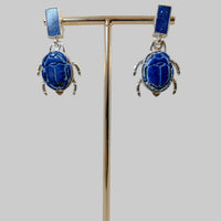 Pair of blue scarab earrings on a gold stand with a wooden base against a gray background