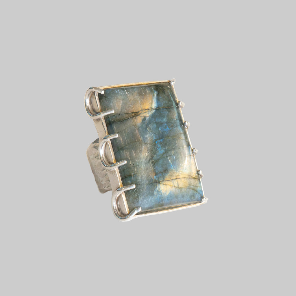 A silver ring featuring a large, rectangular labradorite stone with shimmering blue, gold, and gray flashes, set in a wide band with three prongs.