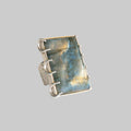 A silver ring featuring a large, rectangular labradorite stone with shimmering blue, gold, and gray flashes, set in a wide band with three prongs.