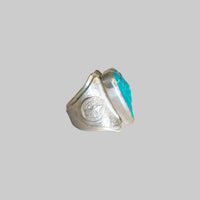 A vintage silver ring with a large, wide band featuring a circular engraved emblem and a prominent turquoise-colored cabochon stone, shown on a plain light background.