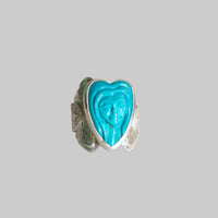 A silver ring featuring a heart-shaped turquoise cabochon with a carved face, flanked by textured green stone inlays on a white background.