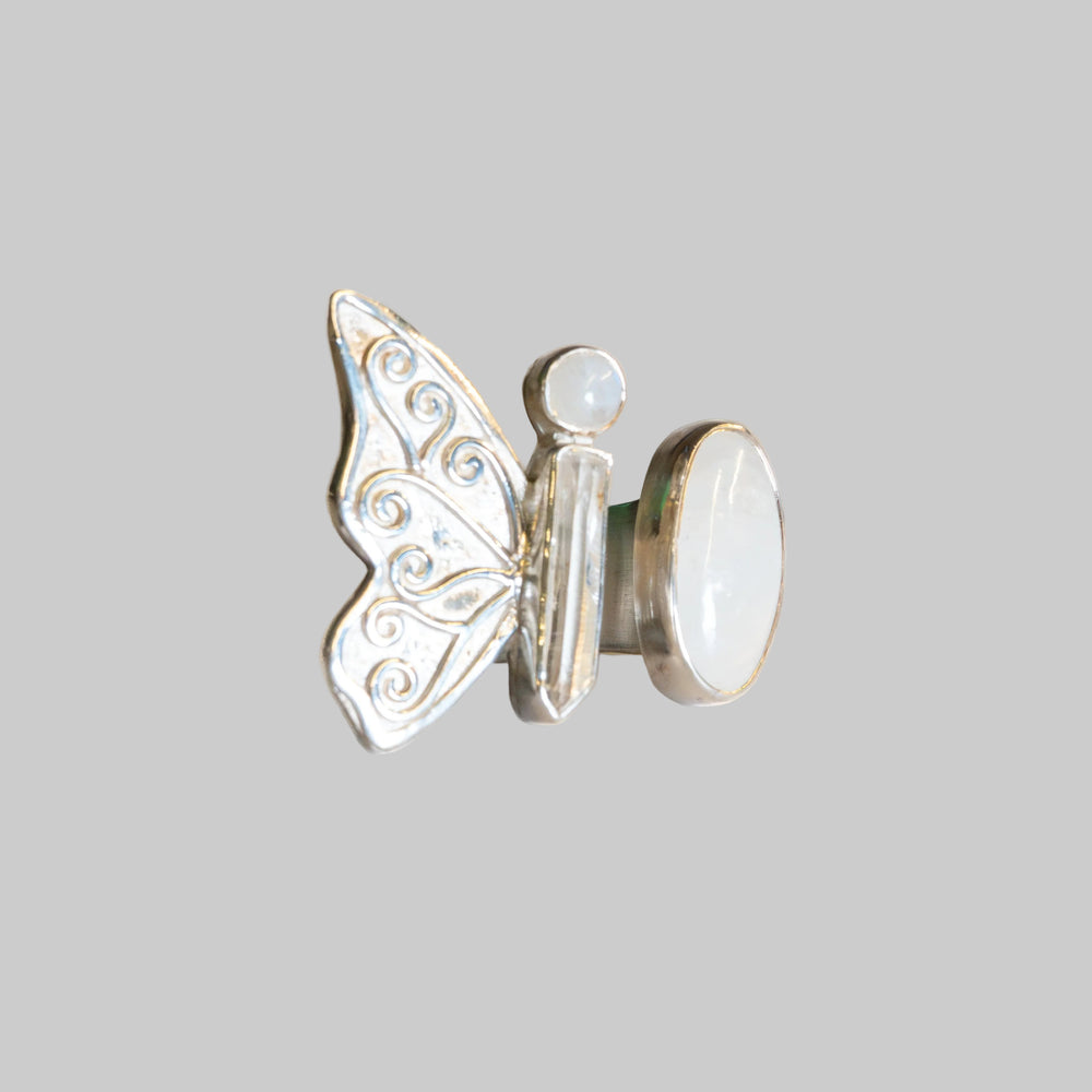 A silver butterfly ring with a white mother-of-pearl oval back and a small round pearl accent, photographed on a neutral cream background.