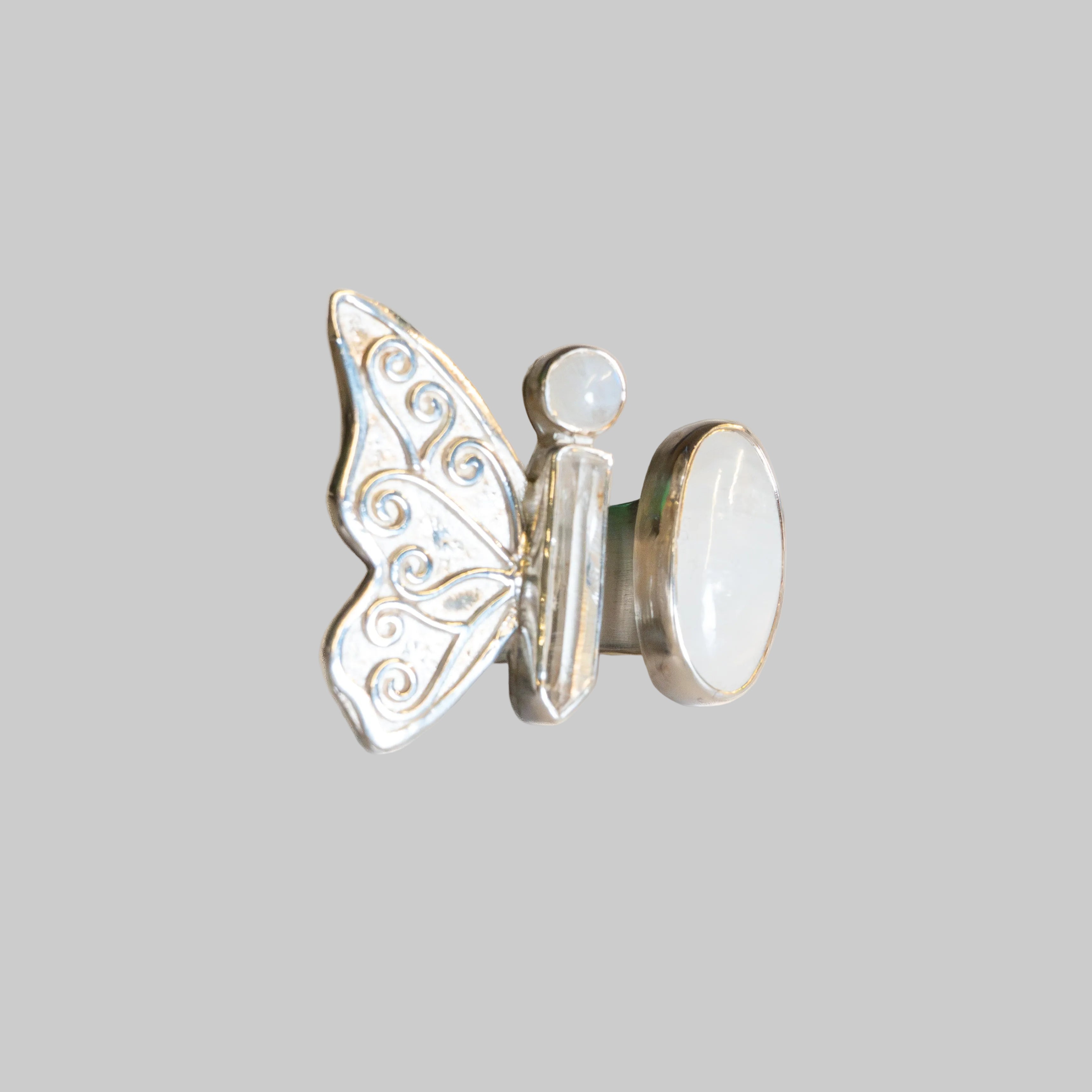 A silver butterfly ring with a white mother-of-pearl oval back and a small round pearl accent, photographed on a neutral cream background.
