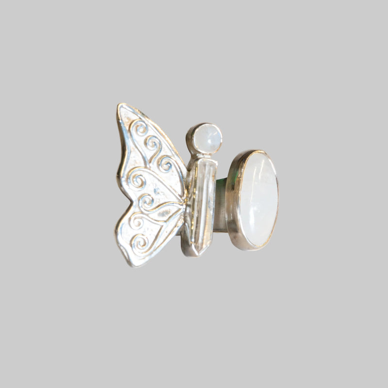 A silver butterfly ring with a white mother-of-pearl oval back and a small round pearl accent, photographed on a neutral cream background.