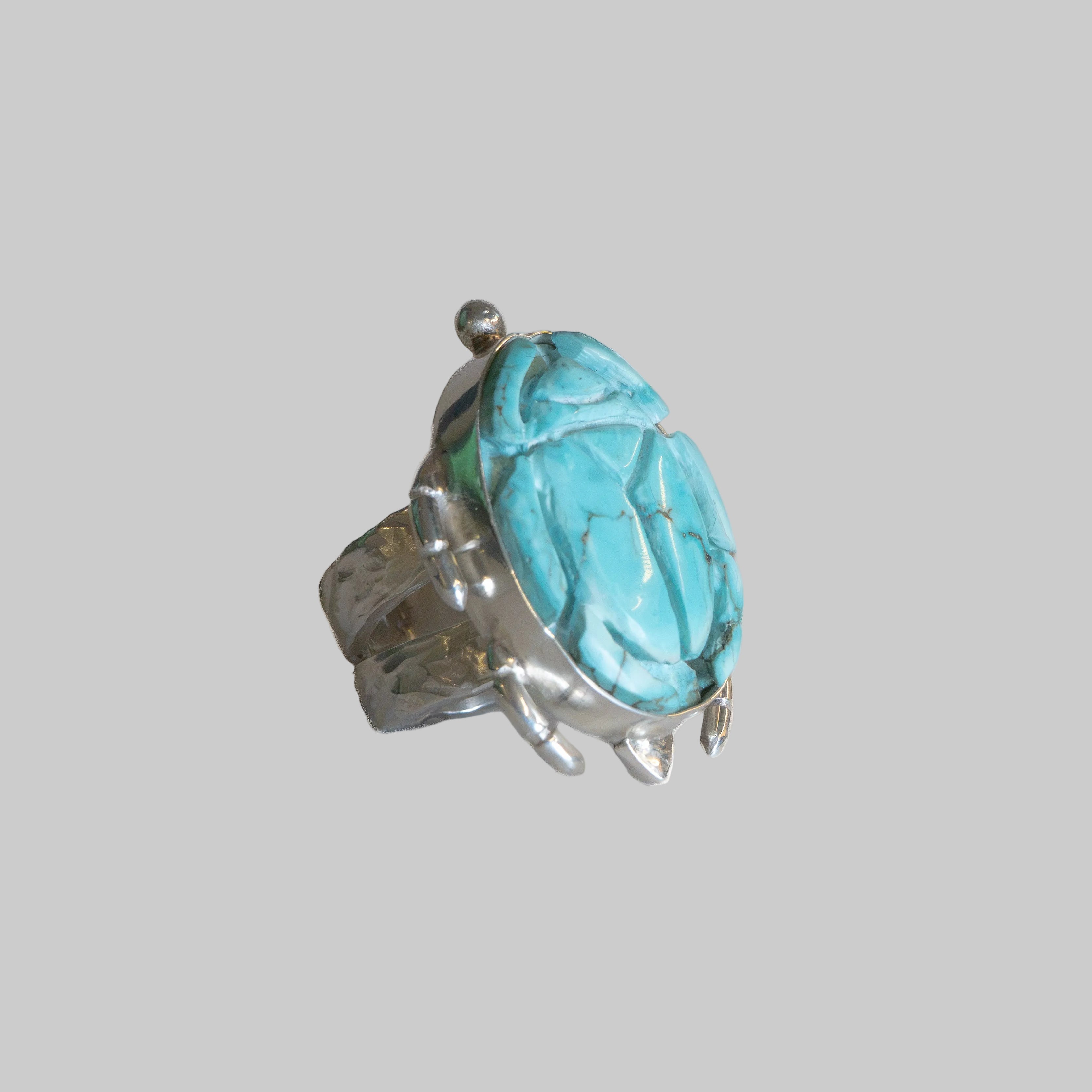 A unique ring designed to look like a beetle, featuring a large turquoise-colored stone on the elytra and a textured silver metal body, set against a plain white background.