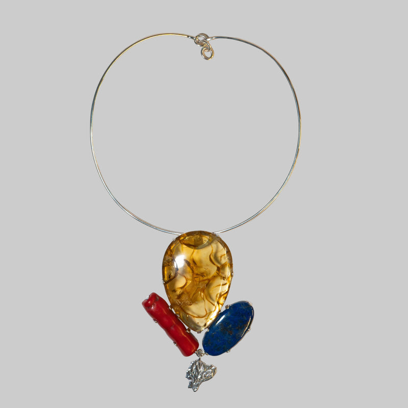 A silver wire circle necklace with three dangling charms: a large teardrop-shaped amber glass piece, a red cylindrical charm, and an oval dark blue glass piece, displayed on a plain white background.