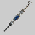 A vintage-style metal bracelet featuring a central oval cabochon with a blue carved figure, flanked by two rectangular links with textured detailing, all connected by dark metal chain links on a plain white background.