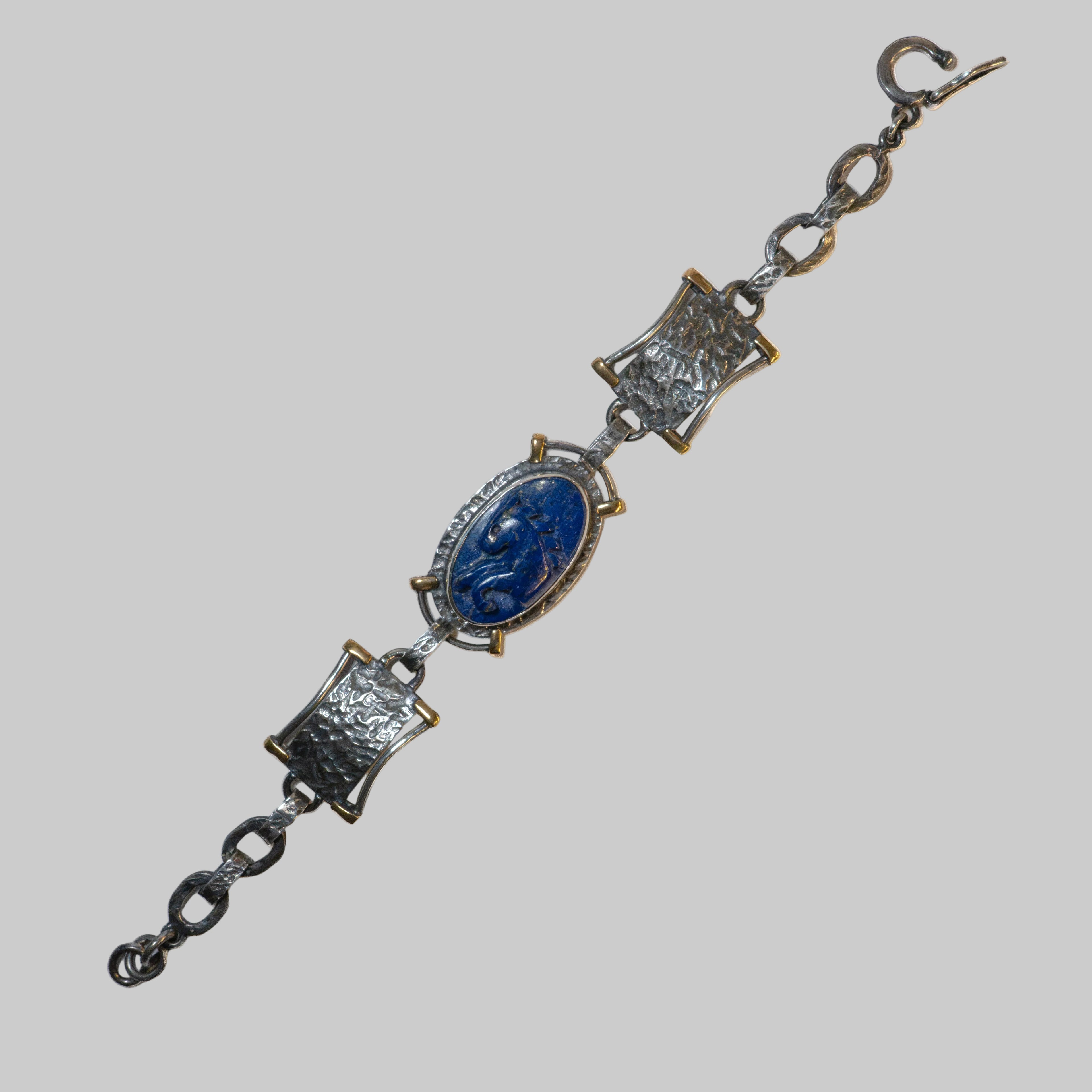 A vintage-style metal bracelet featuring a central oval cabochon with a blue carved figure, flanked by two rectangular links with textured detailing, all connected by dark metal chain links on a plain white background.