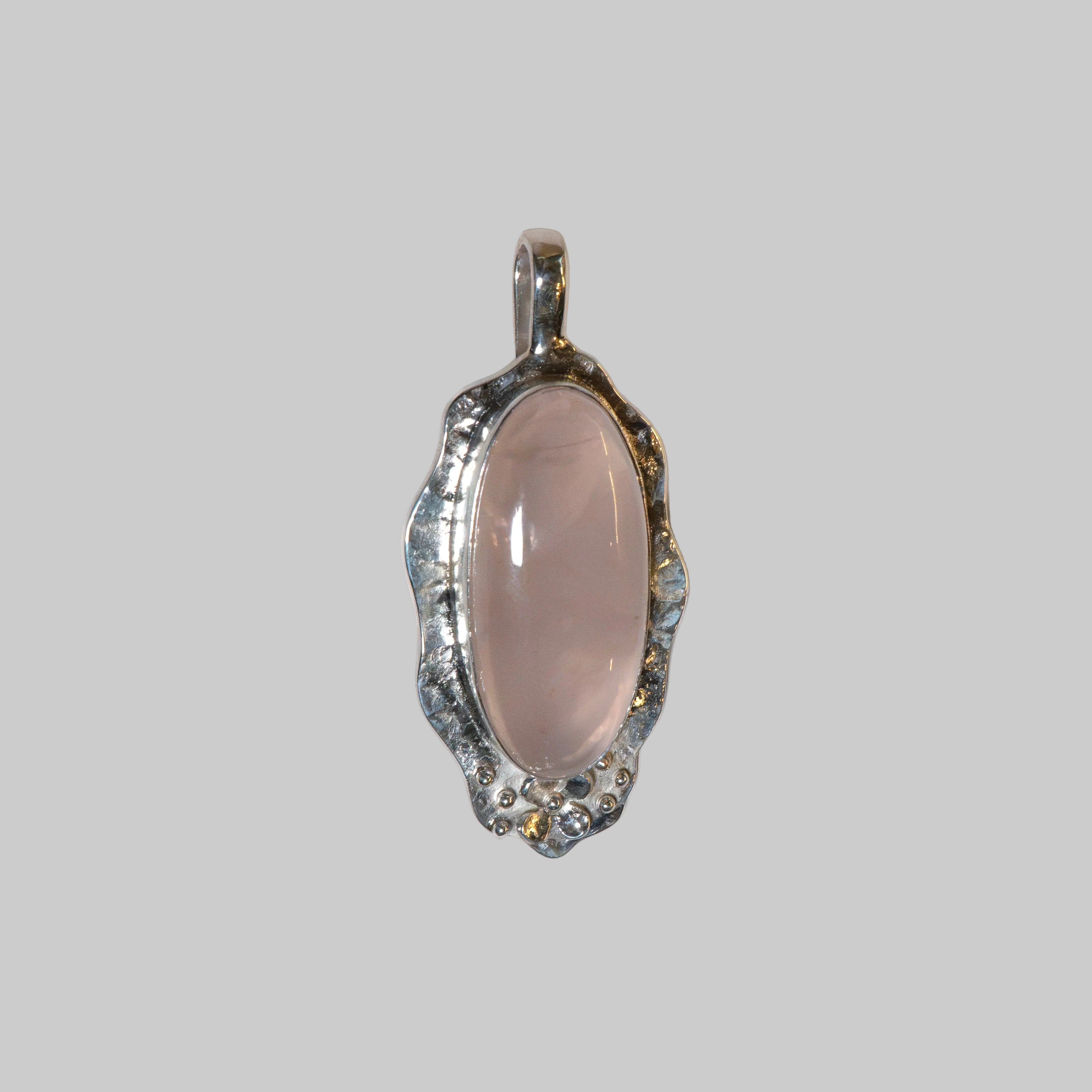 An oval pendant featuring a smooth, pale pink cabochon stone set in a textured, silver-toned bezel with small clear stones along the scalloped edge, shown on a plain white background.