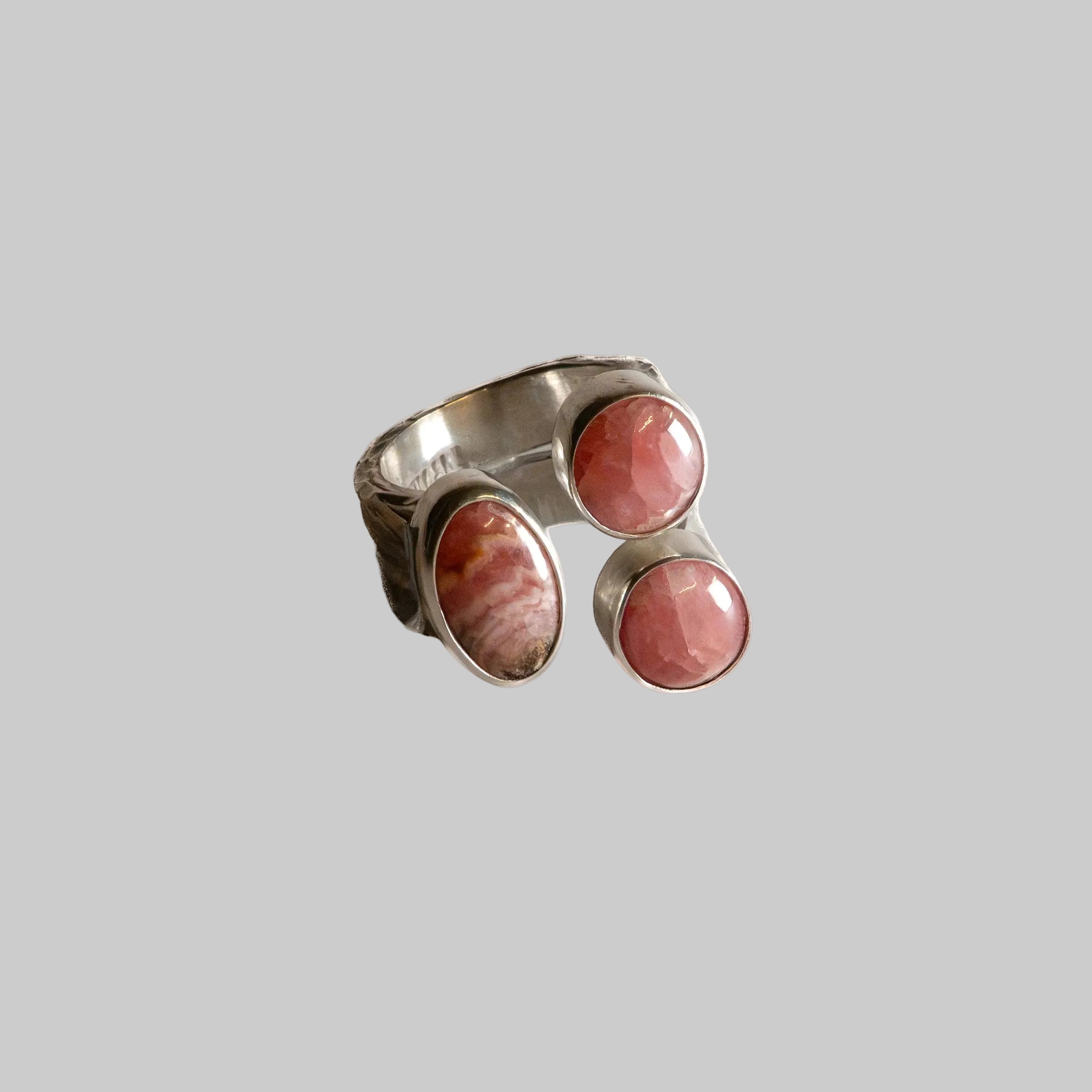 A silver-toned ring featuring three oval pinkish-red stone cabochons set in a wide band, photographed on a plain off-white background.