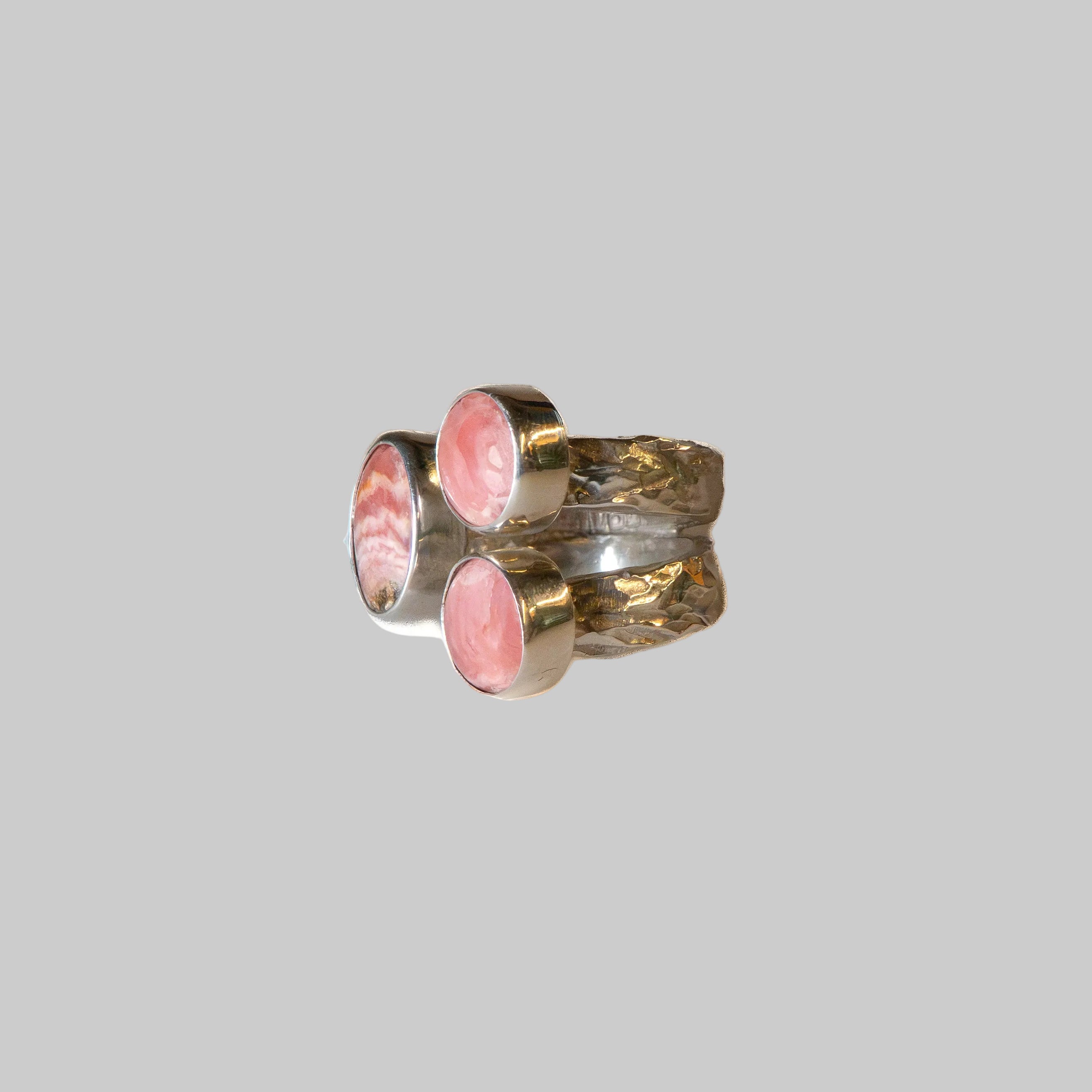A unique textured silver ring featuring two oval pink mother-of-pearl inlays, photographed on a clean, light beige gradient background.